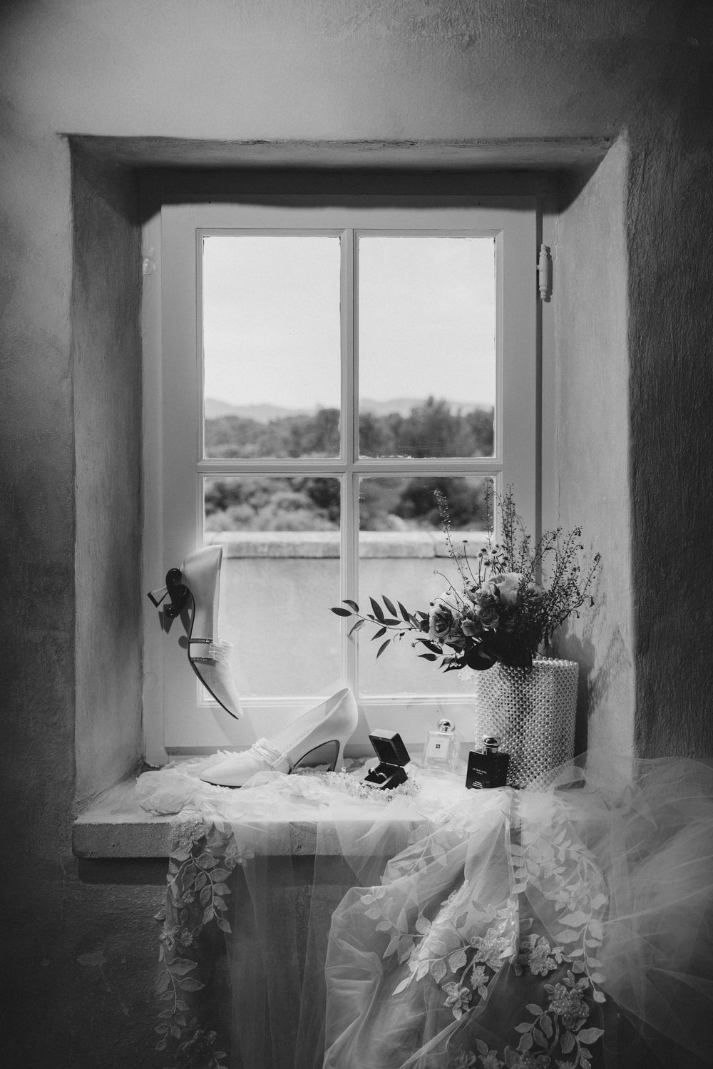 A black and white photograph of a rustic window sill with bridal shoes and floral arrangements.