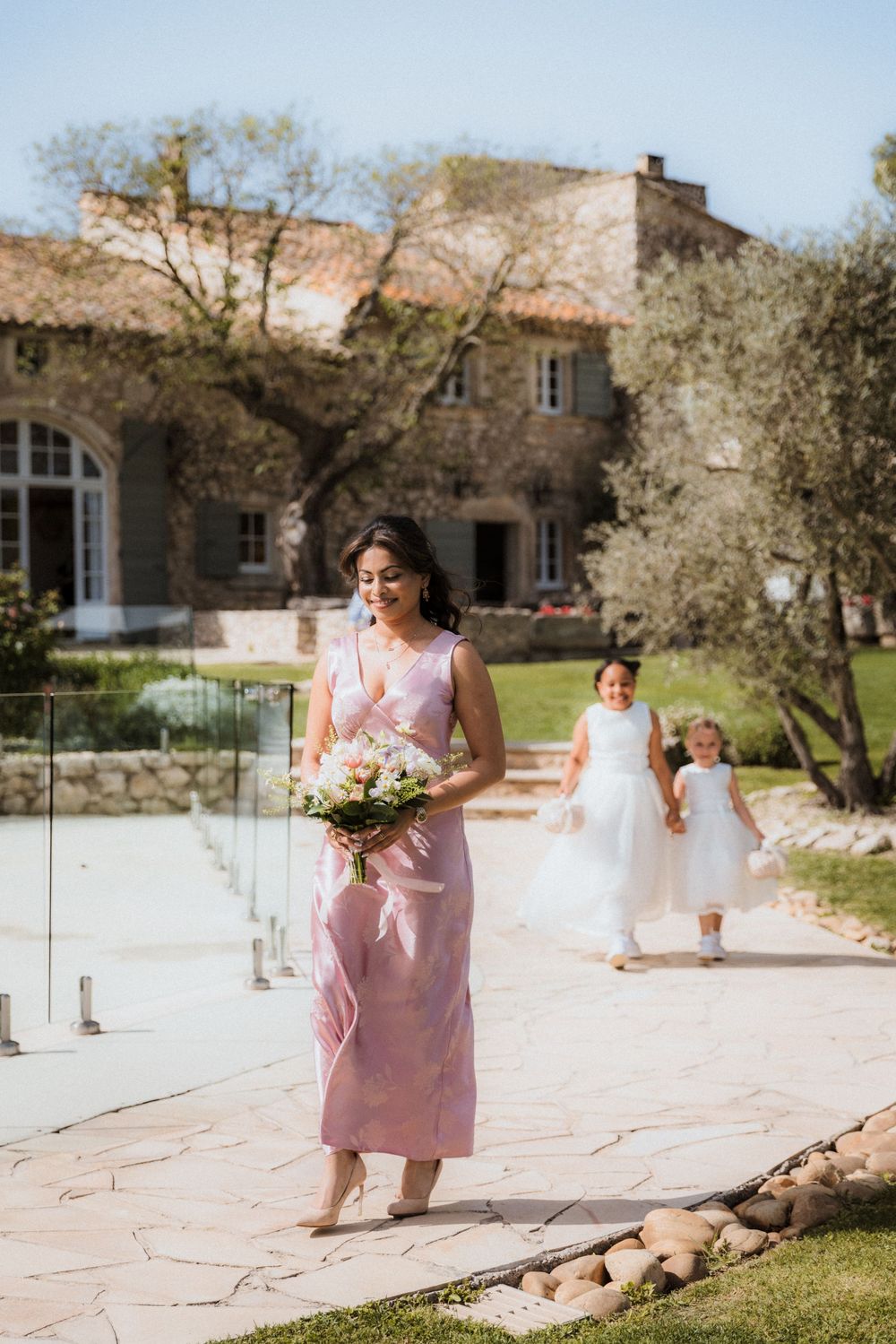 Bridesmaids in pink dresses walk along a stone path carrying bouquets near a Mediterranean-style building.