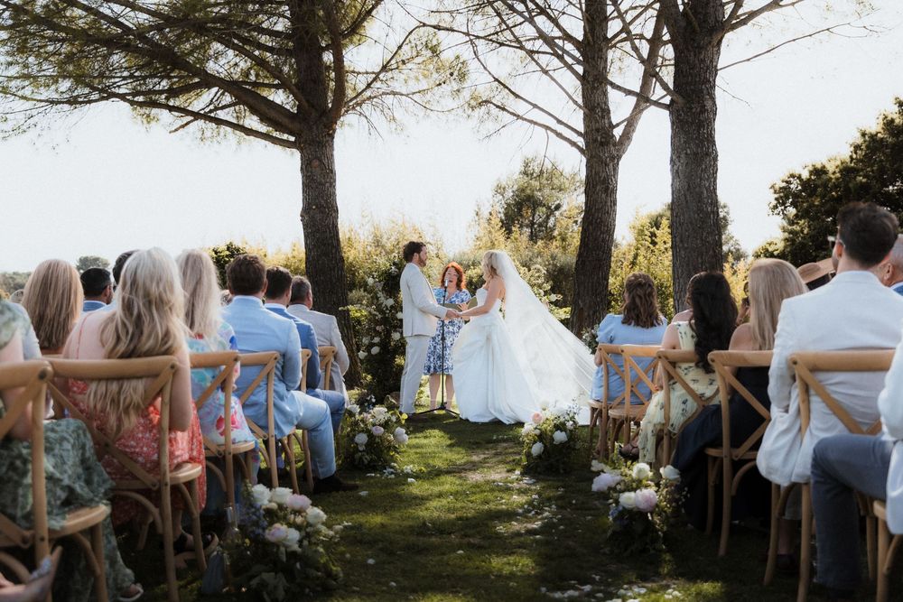Outdoor wedding ceremony under tall trees with guests seated on wooden chairs.