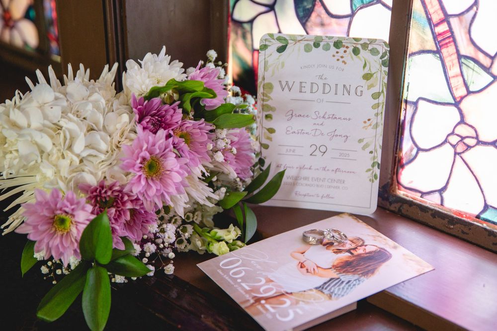 Wedding invitation displayed next to a floral arrangement of white and pink flowers by a stained glass window.