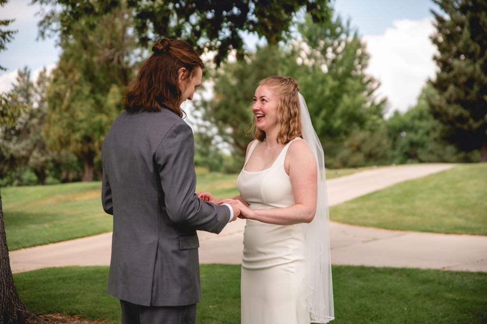 Two people exchange rings during an outdoor wedding ceremony on a tree-lined path in a grassy area.
