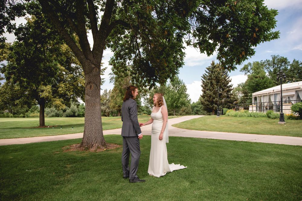 A couple stands together on a manicured lawn with large shade trees and a building visible in the background.