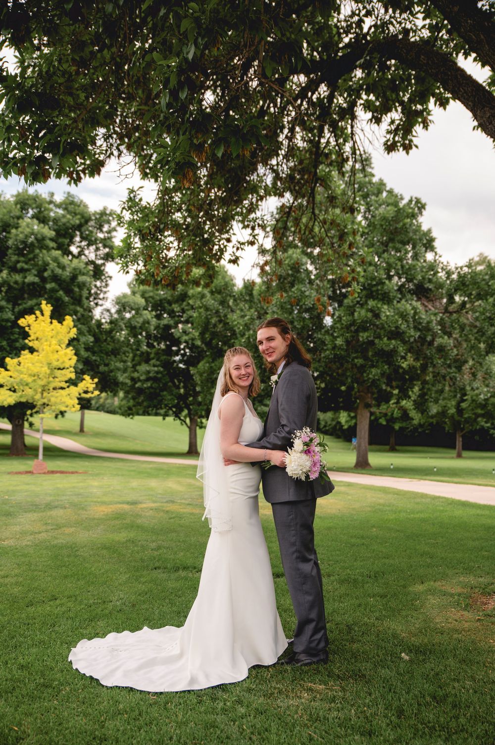 A couple in wedding attire poses together on a lush green lawn bordered by trees on their special day.