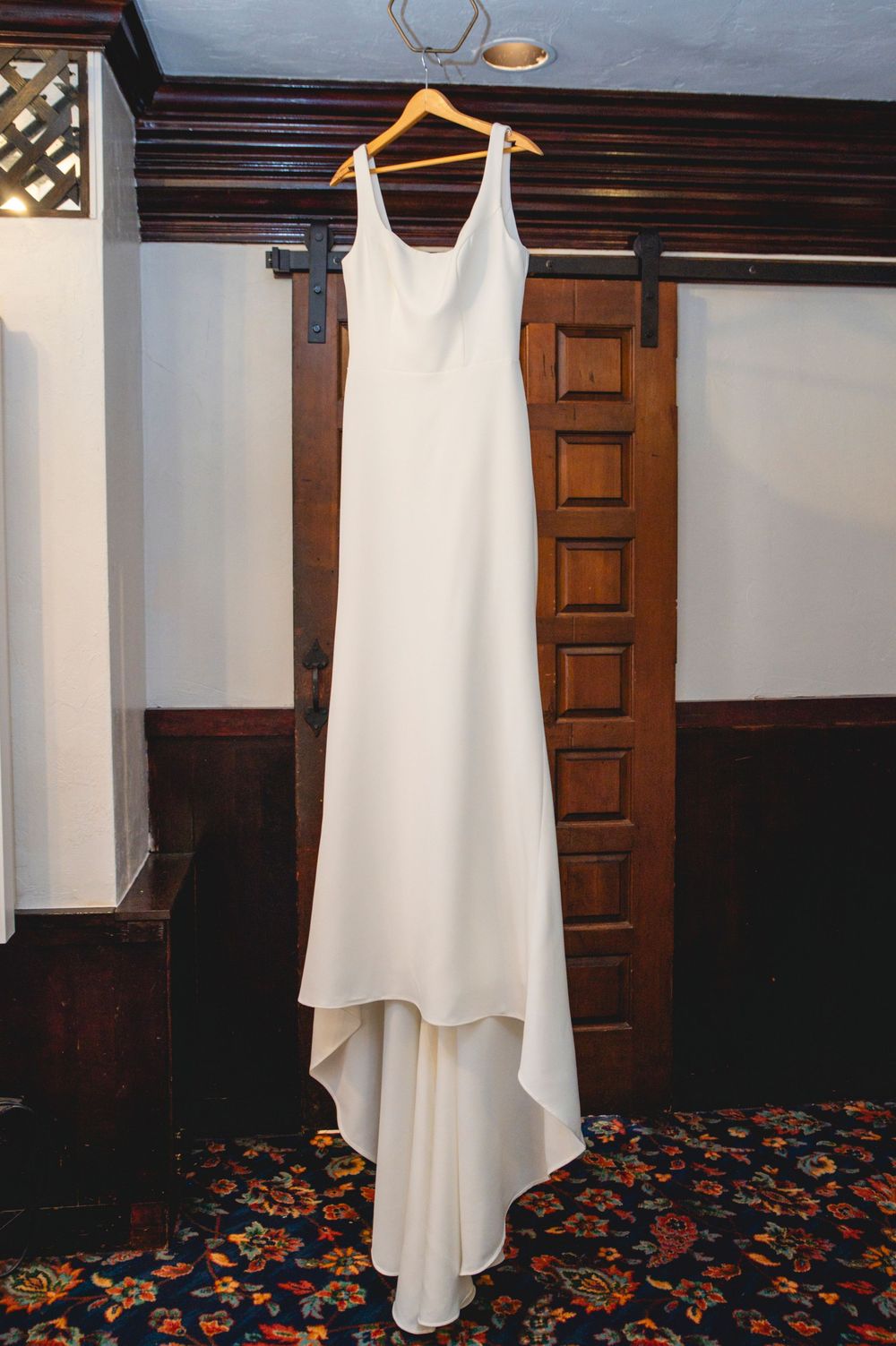 White wedding dress hanging on wooden hanger against wood-paneled wall with floral carpeting below.