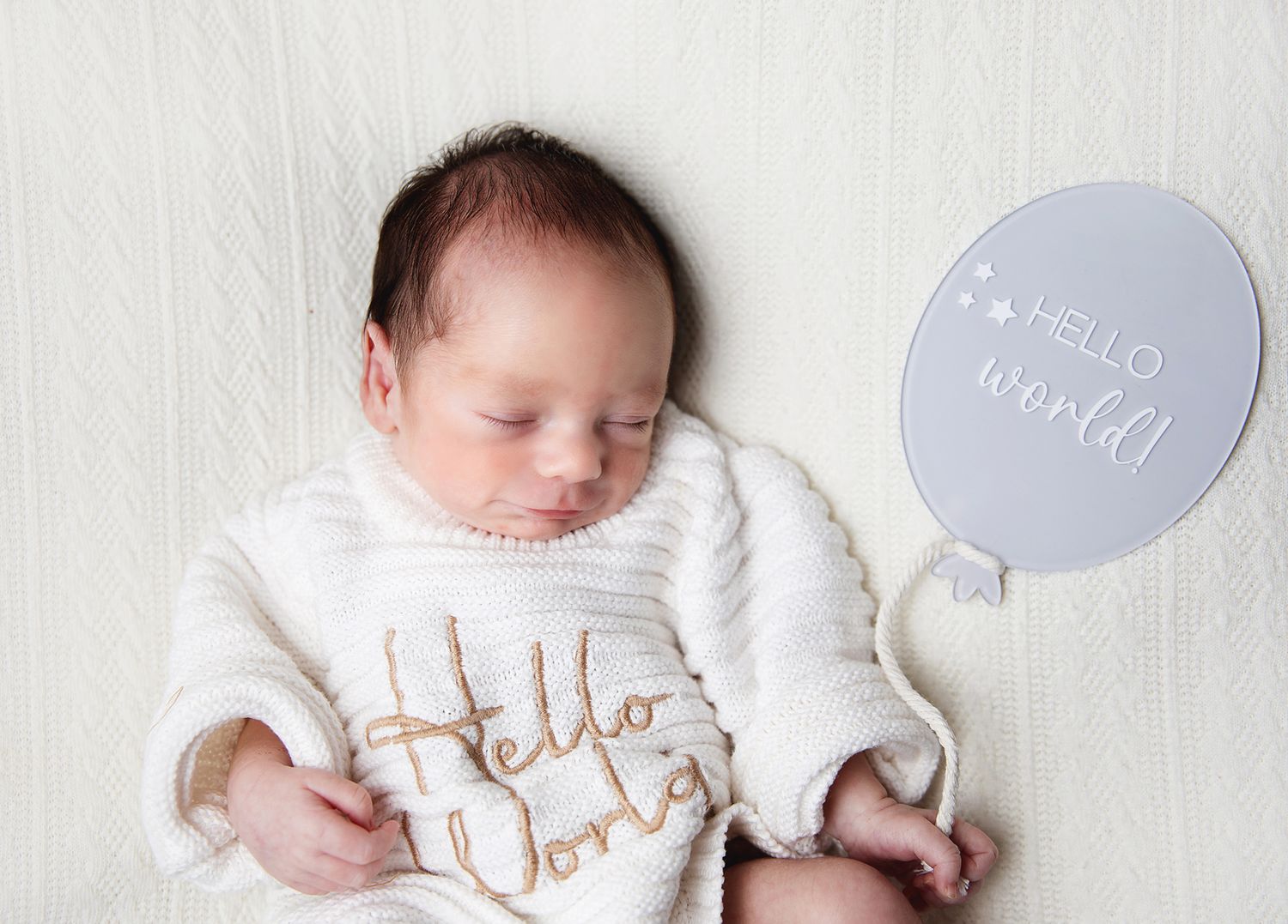A sleeping newborn in a white 'Hello World' onesie lies peacefully on a soft white blanket. During a newborn session in South East London