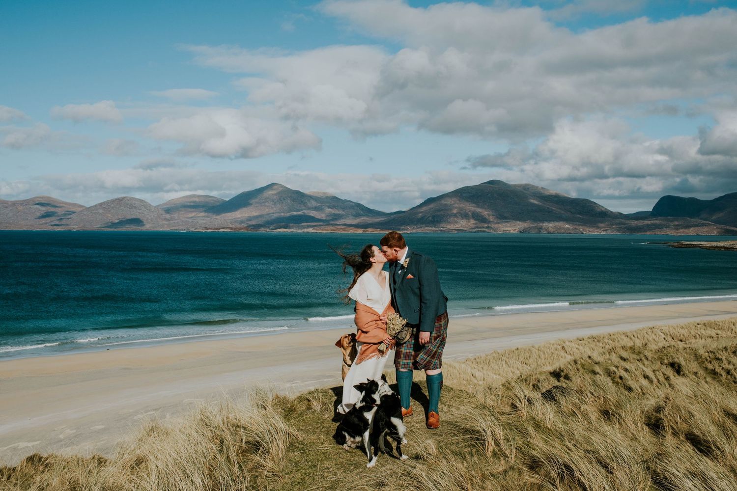 A couple in wedding attire stand overlooking a scenic Scottish loch with dramatic clouds and mountains in the background.