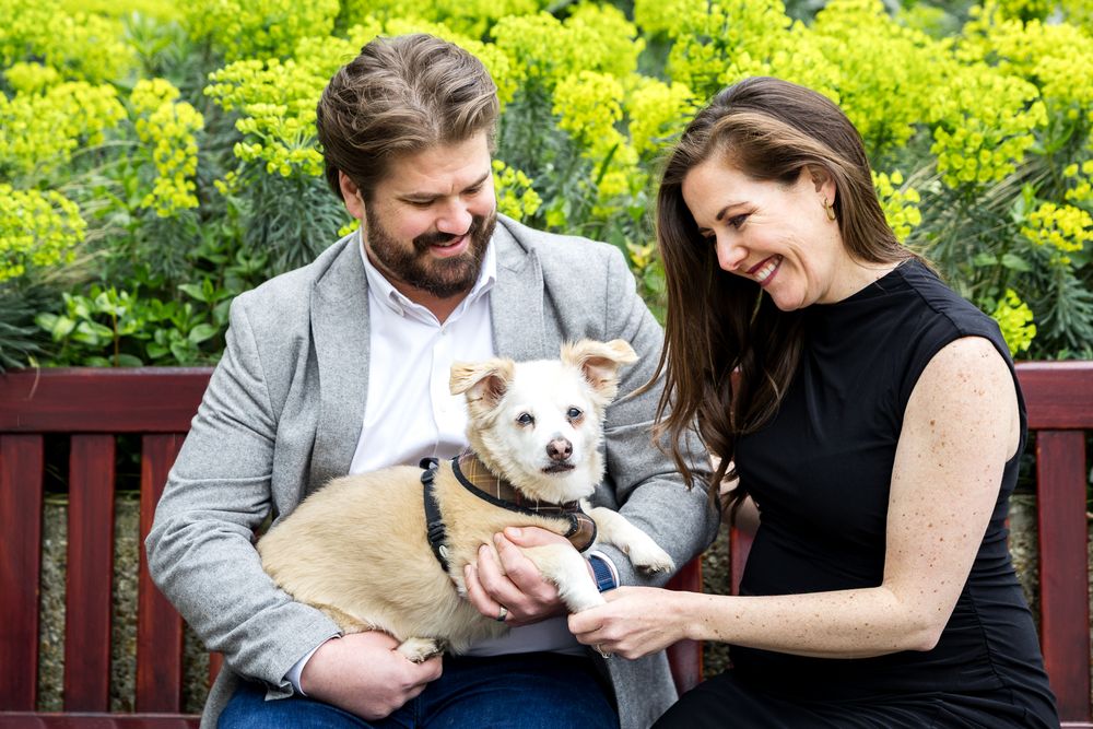 husband and wife sitting on bench in front of green flowers with their cream coloured dog