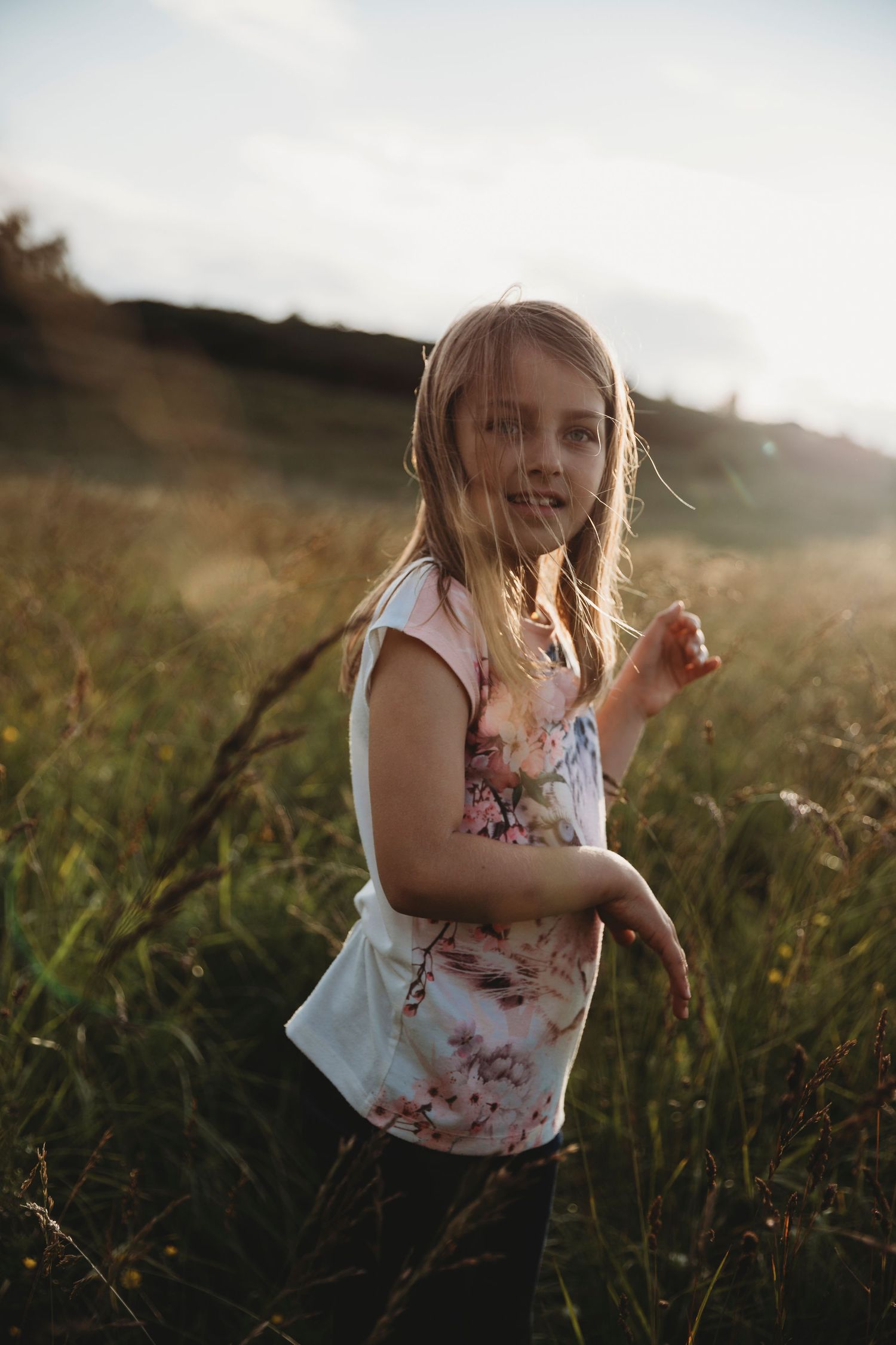 Child wearing a floral sleeveless top plays in a grassy field at golden hour with sunlight glowing behind.