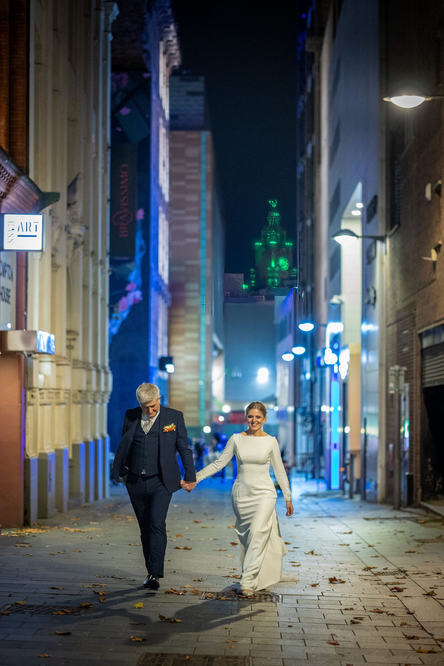 bride and groom walk through a street in Liverpool City Centre the liver building lit up green in the distance. Stuart Morris Photography