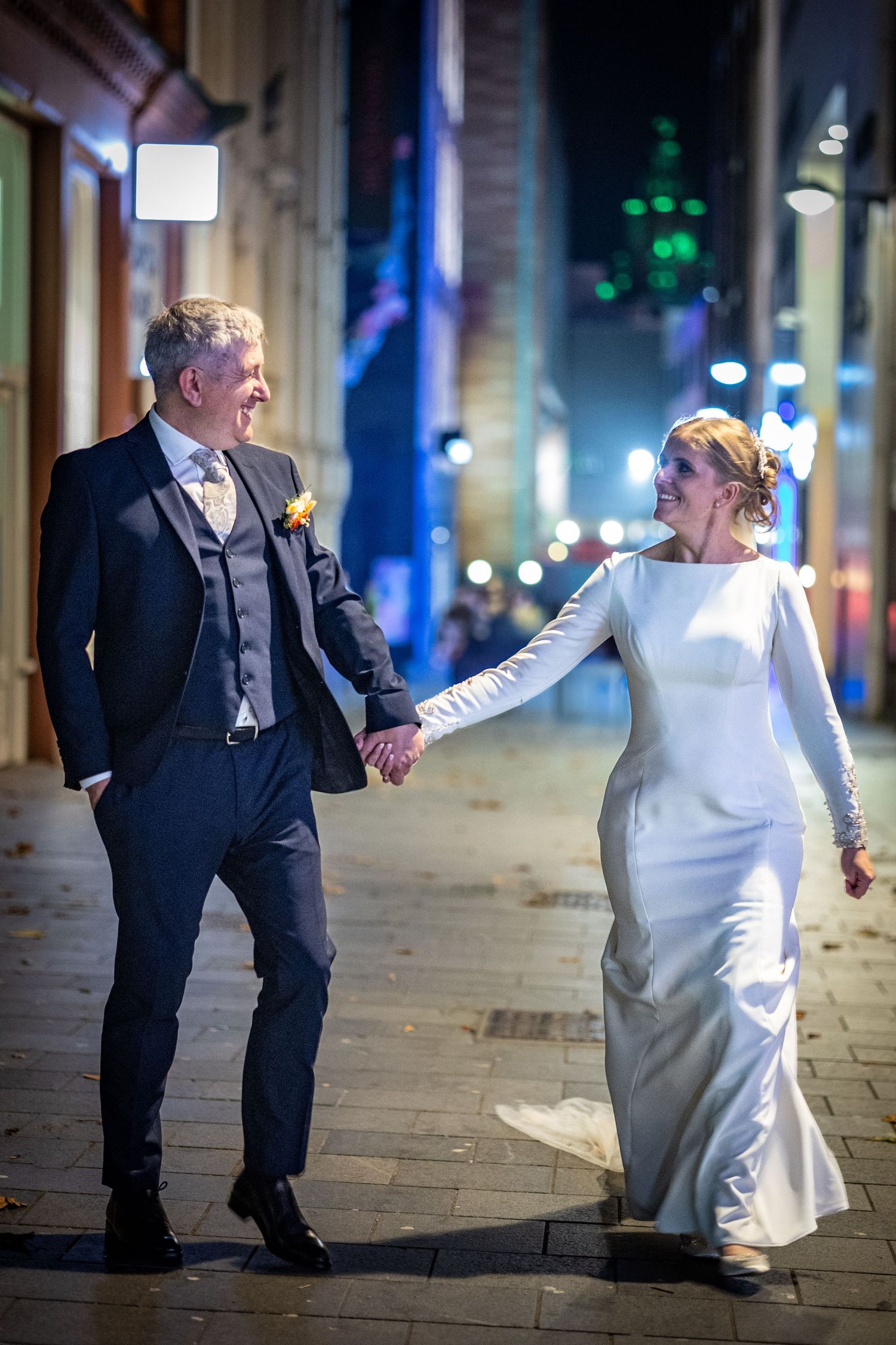 bride and groom holding hands at night walk through liverpool city centre with the liver building in the background behind them.