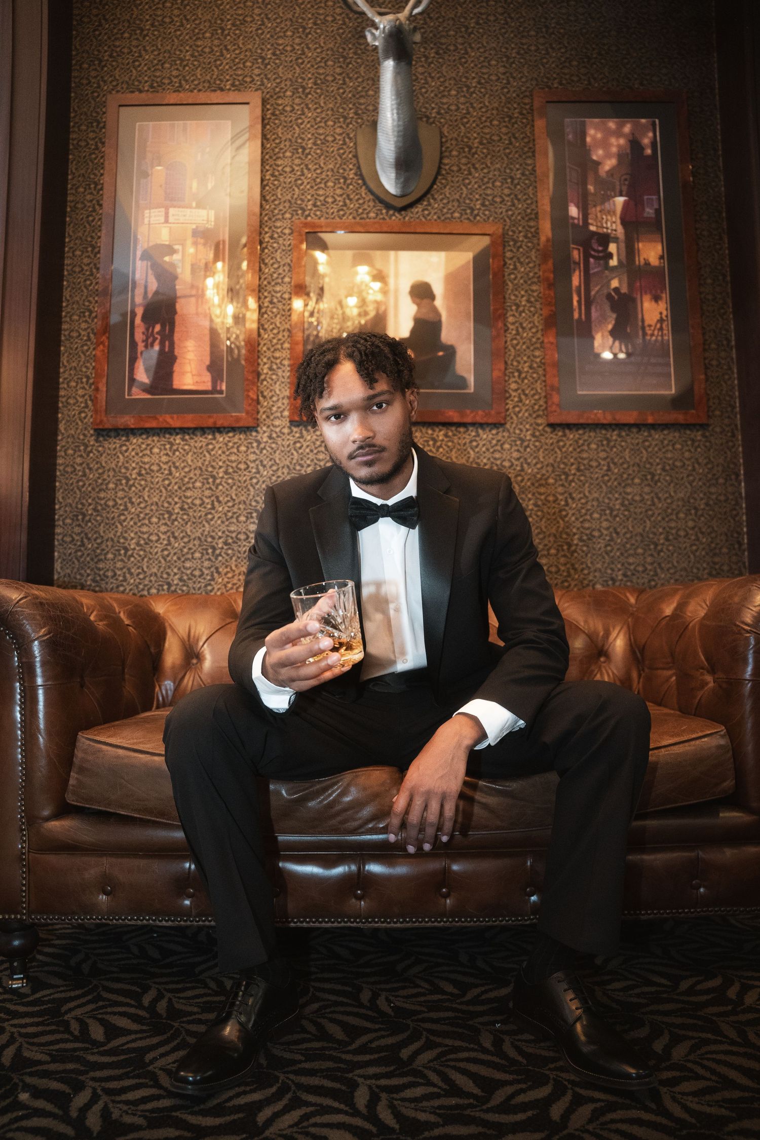 A person in a tuxedo sits on a brown leather couch in an elegant room with framed photos on textured wallpaper.