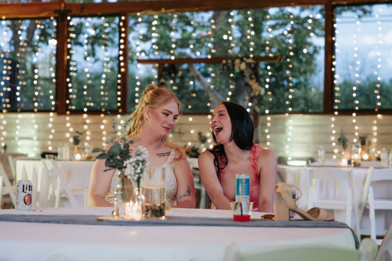 Wedding guests chat and laugh around elegant white-clothed tables at an outdoor reception under string lights.