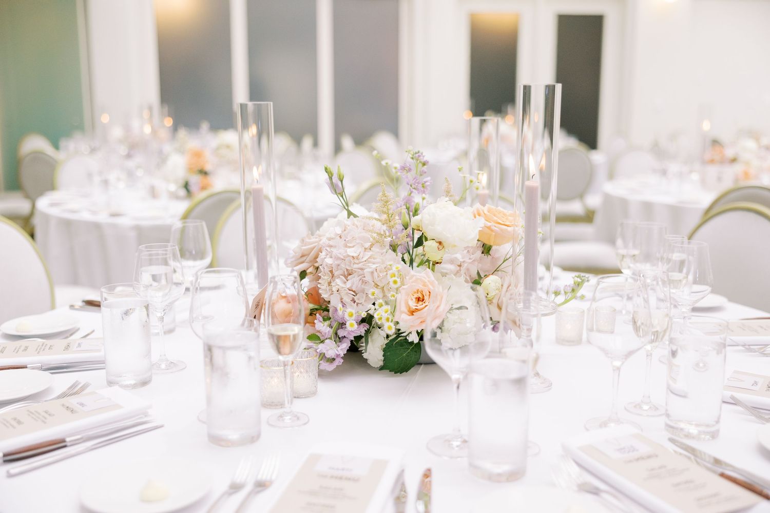 Elegant wedding reception table setting with blush floral centerpiece, white linens, and crystal glassware.