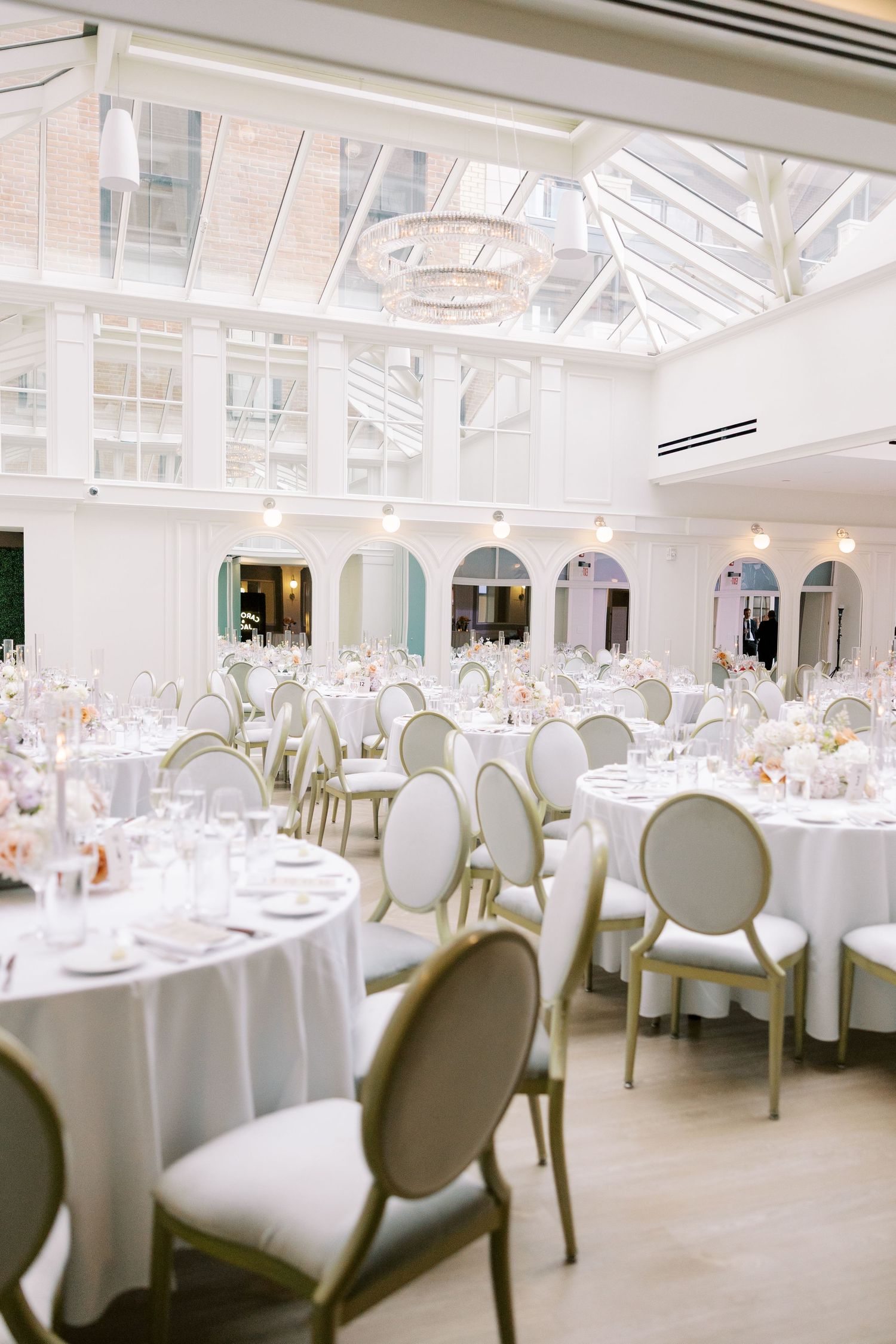 Elegant white wedding venue with glass ceiling, crystal chandelier and round tables set with formal white linens and chairs.
