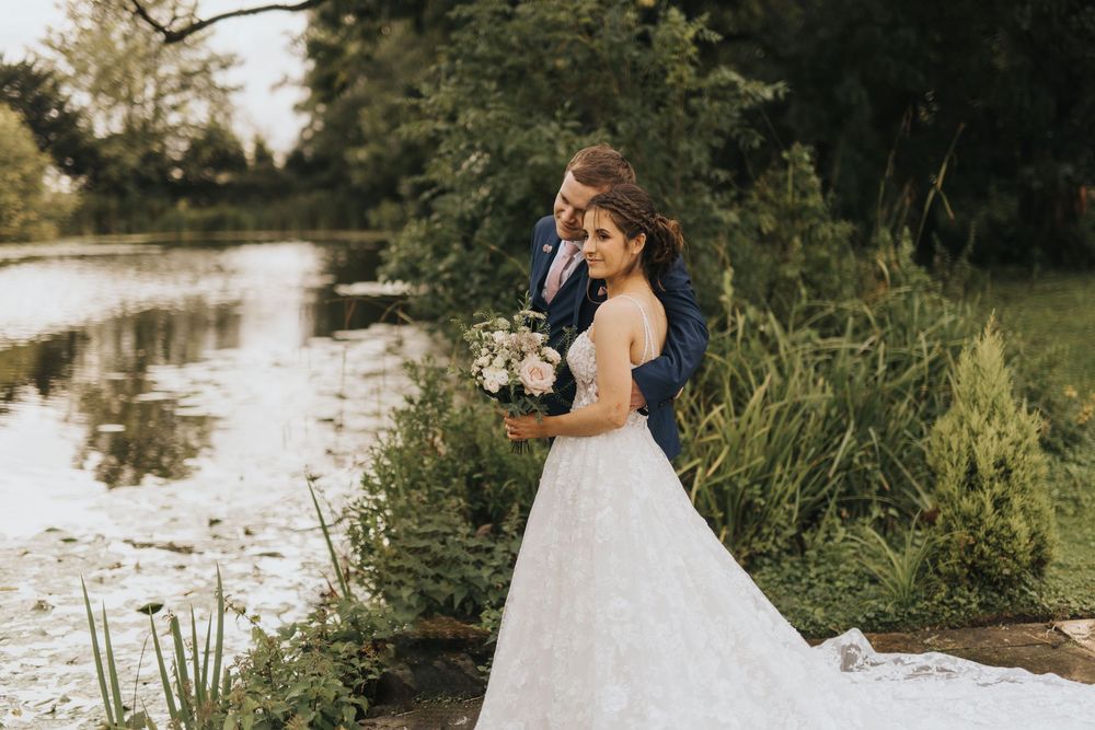 A romantic couple embraces by a tranquil river during their outdoor wedding photoshoot at sunset at Cave Castle.
