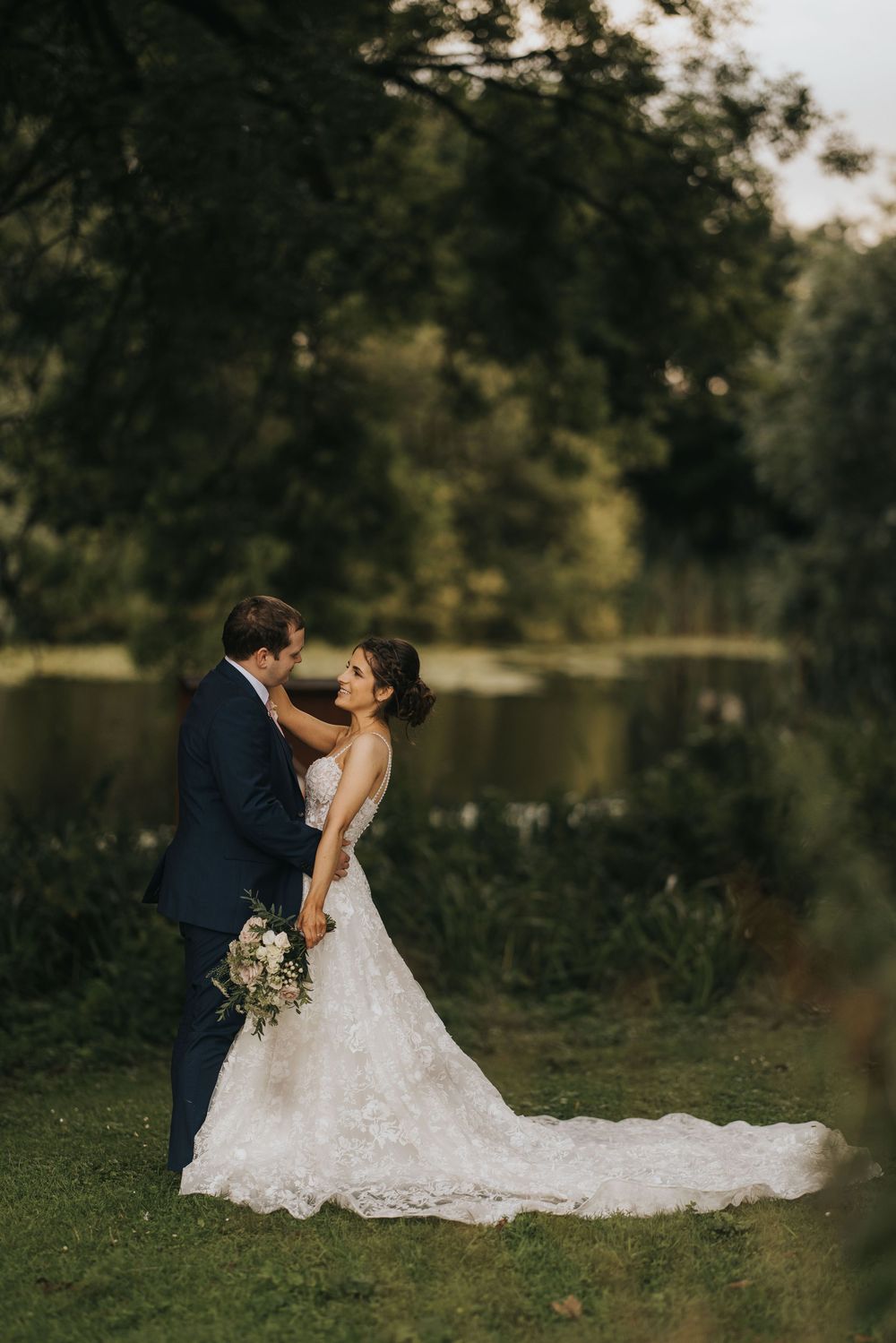 A romantic wedding couple embraces by a serene lake at sunset, with flowing lace gown and navy suit at Cave Castle.