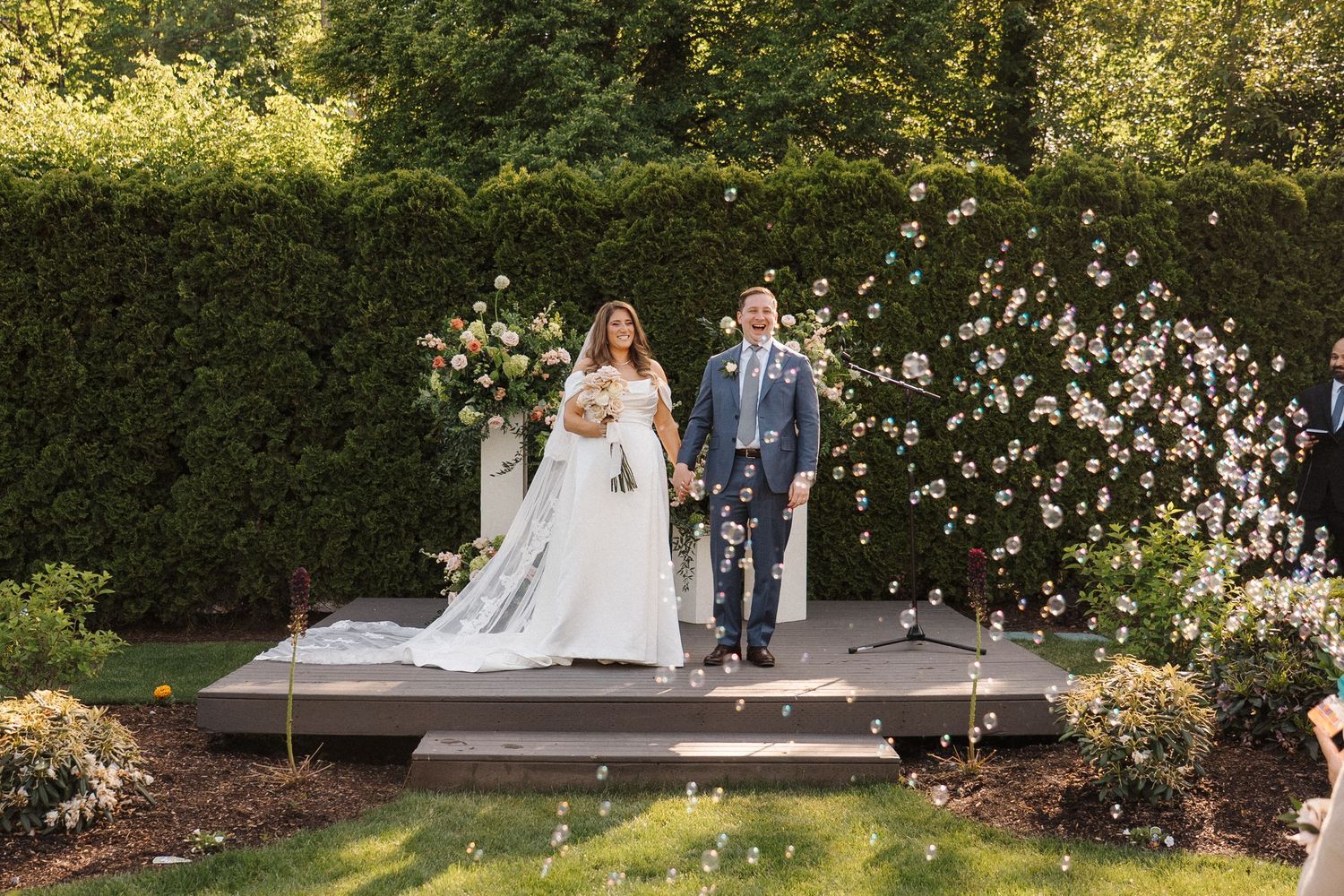 Couple sits together at a garden wedding ceremony venue surrounded by lush greenery and white flowers.