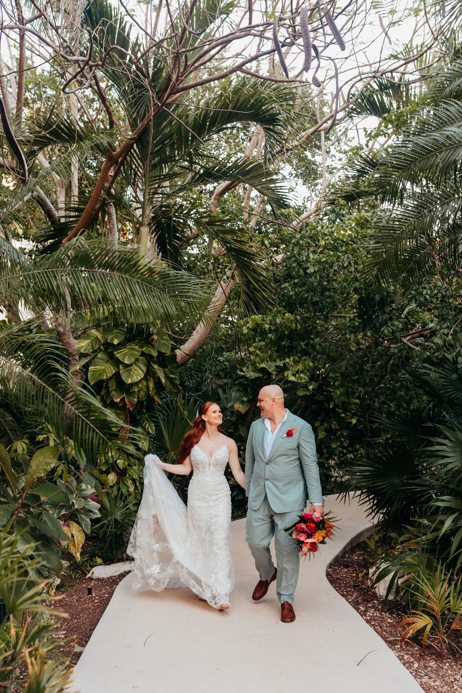 A newlywed couple walking along a tropical garden path surrounded by palm trees and lush greenery.