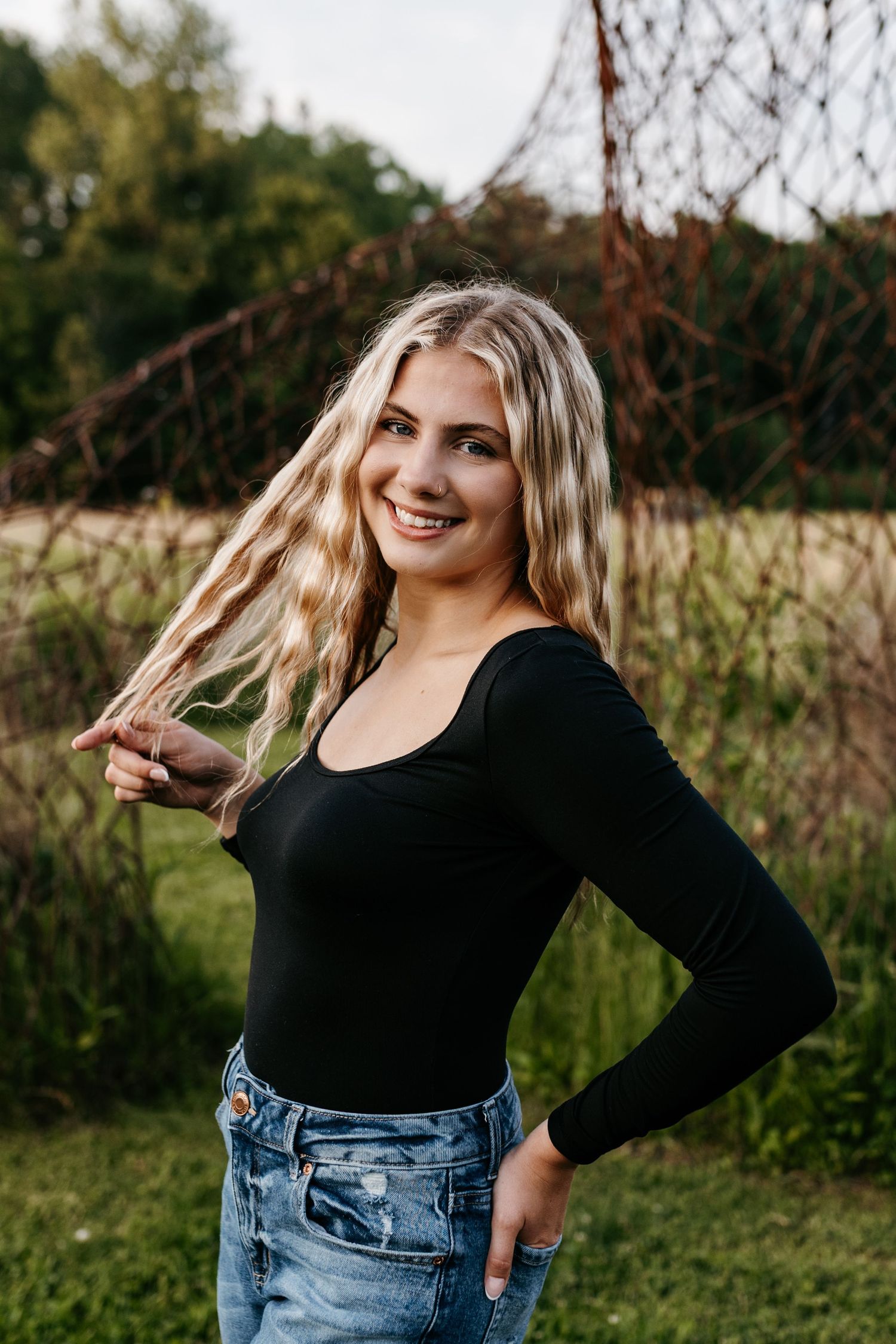 Blonde woman wearing black long sleeve top and blue jeans poses playfully outdoors in natural setting.