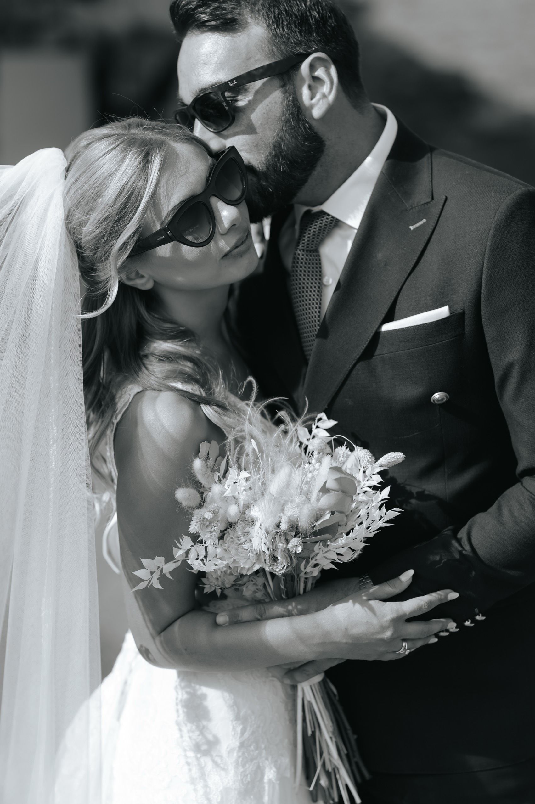 A romantic black and white wedding photograph of a couple embracing, with the bride holding a delicate floral bouquet.