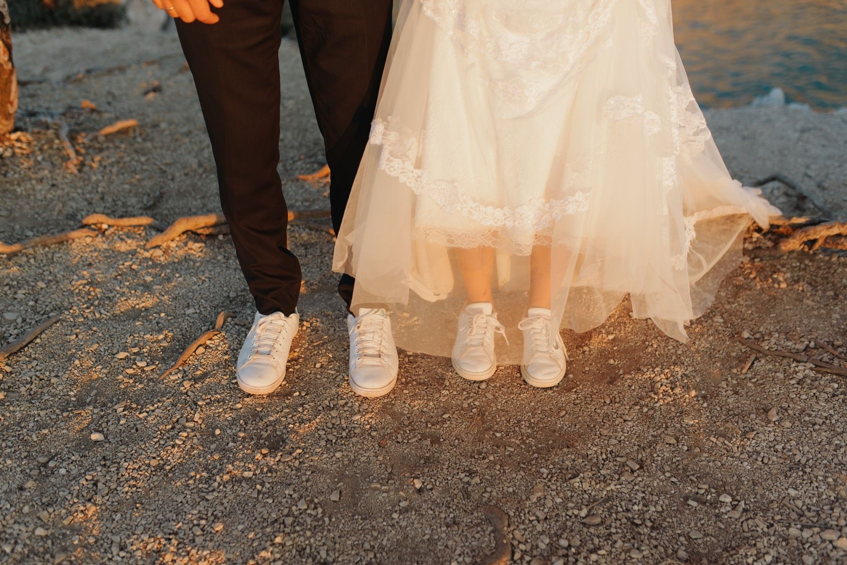 Romantic couple's feet with wedding shoes and dress shown in close-up on sandy ground during sunset.