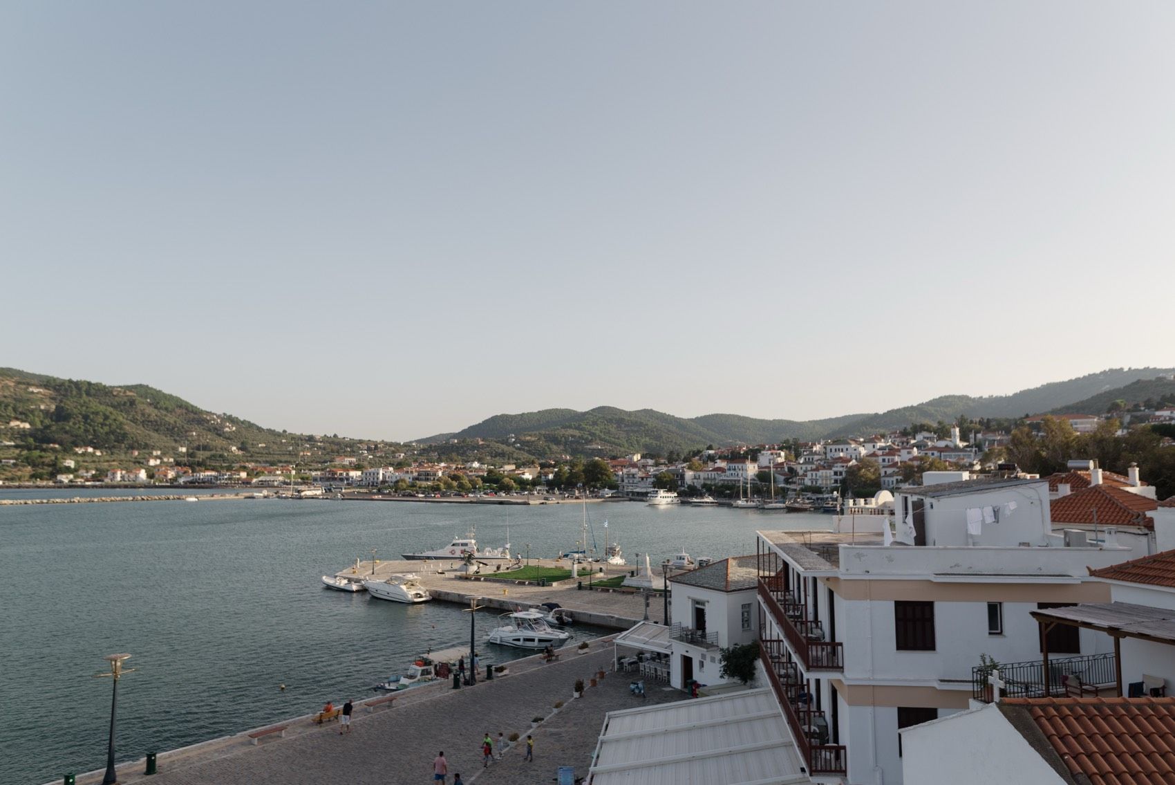 Panoramic view of a coastal Mediterranean village with white buildings nestled along the waterfront and mountains in background.