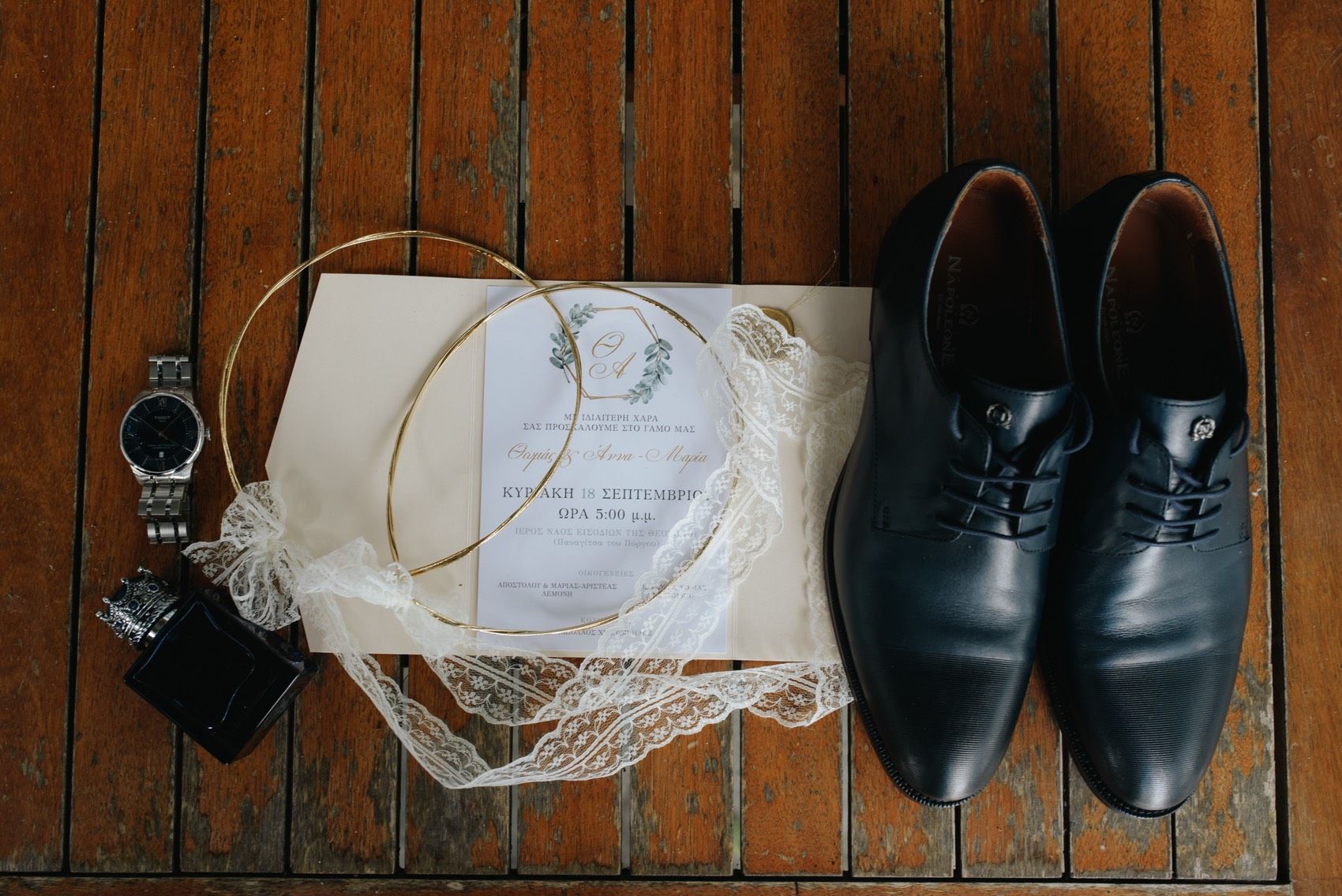 Wedding accessories including black leather dress shoes, invitation, and watch arranged on wooden floor.