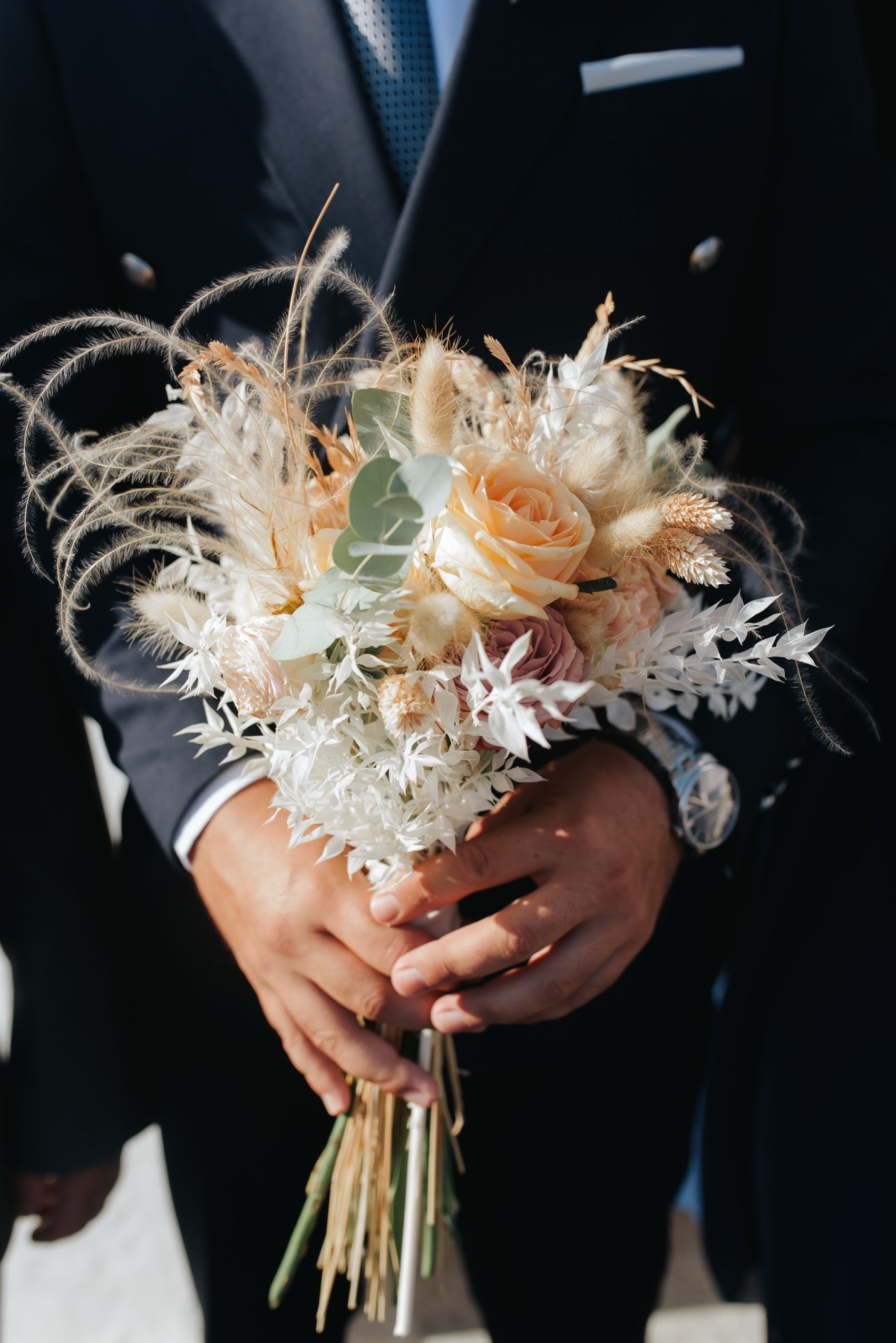 A delicate bridal bouquet of peach roses and wispy dried flowers held by hands in a dark suit.