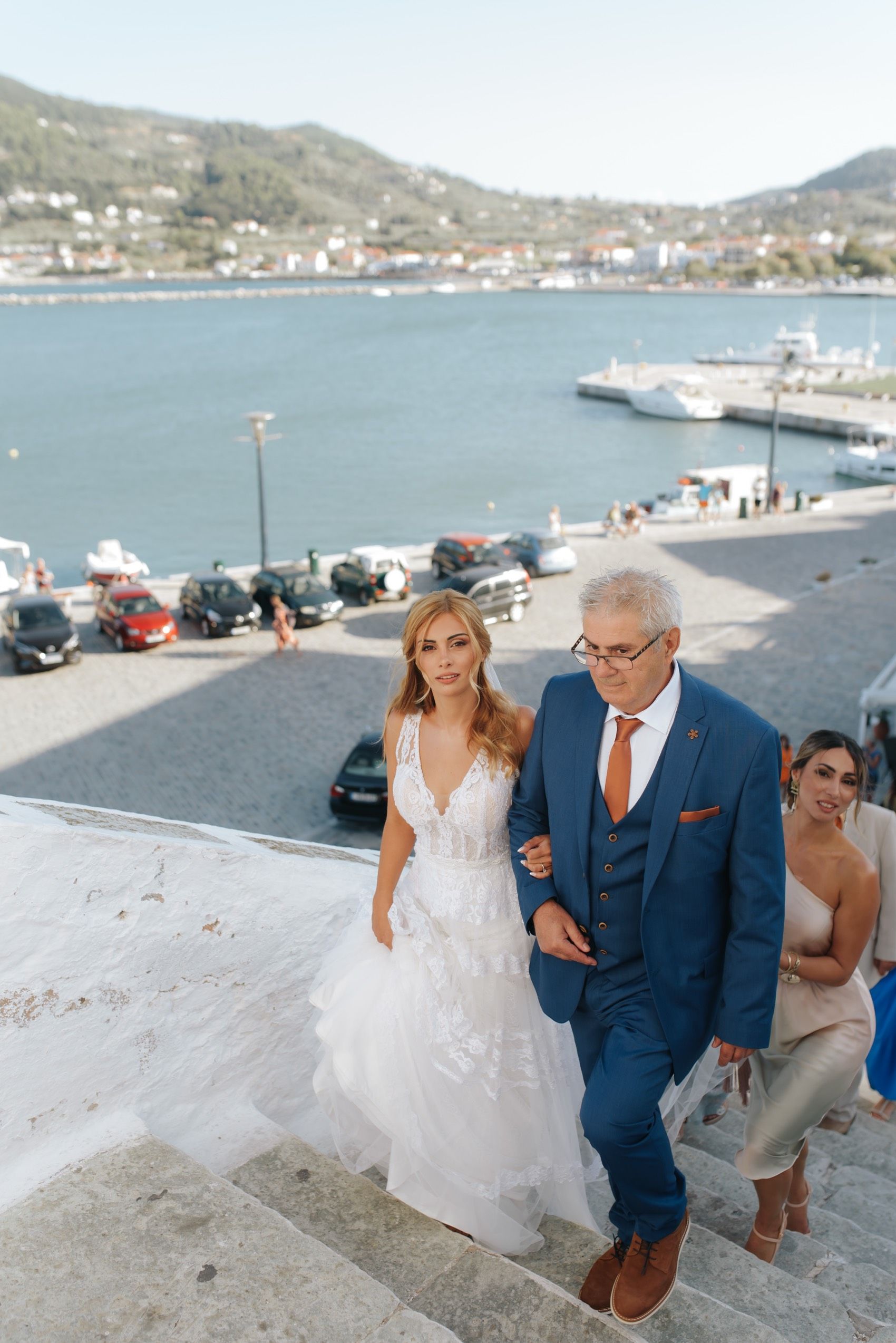 People in wedding attire walk along a scenic waterfront promenade with mountains and boats in the background.
