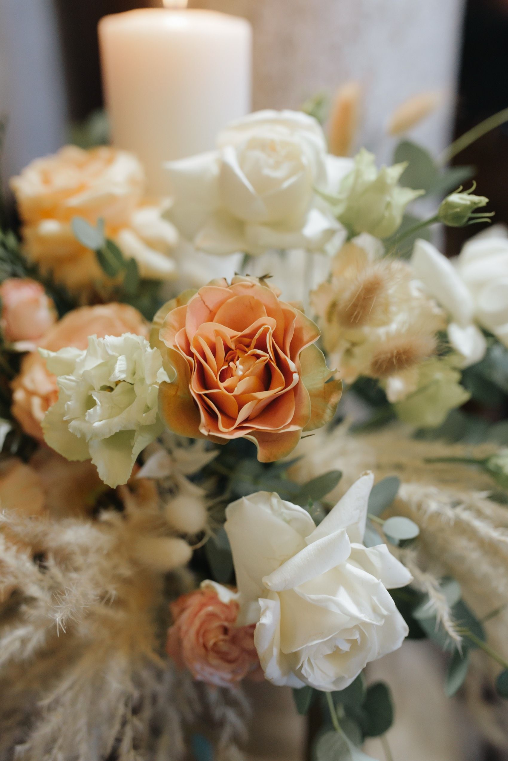 Elegant floral arrangement featuring peach roses, white roses, and dried pampas grass with a white candle centerpiece.