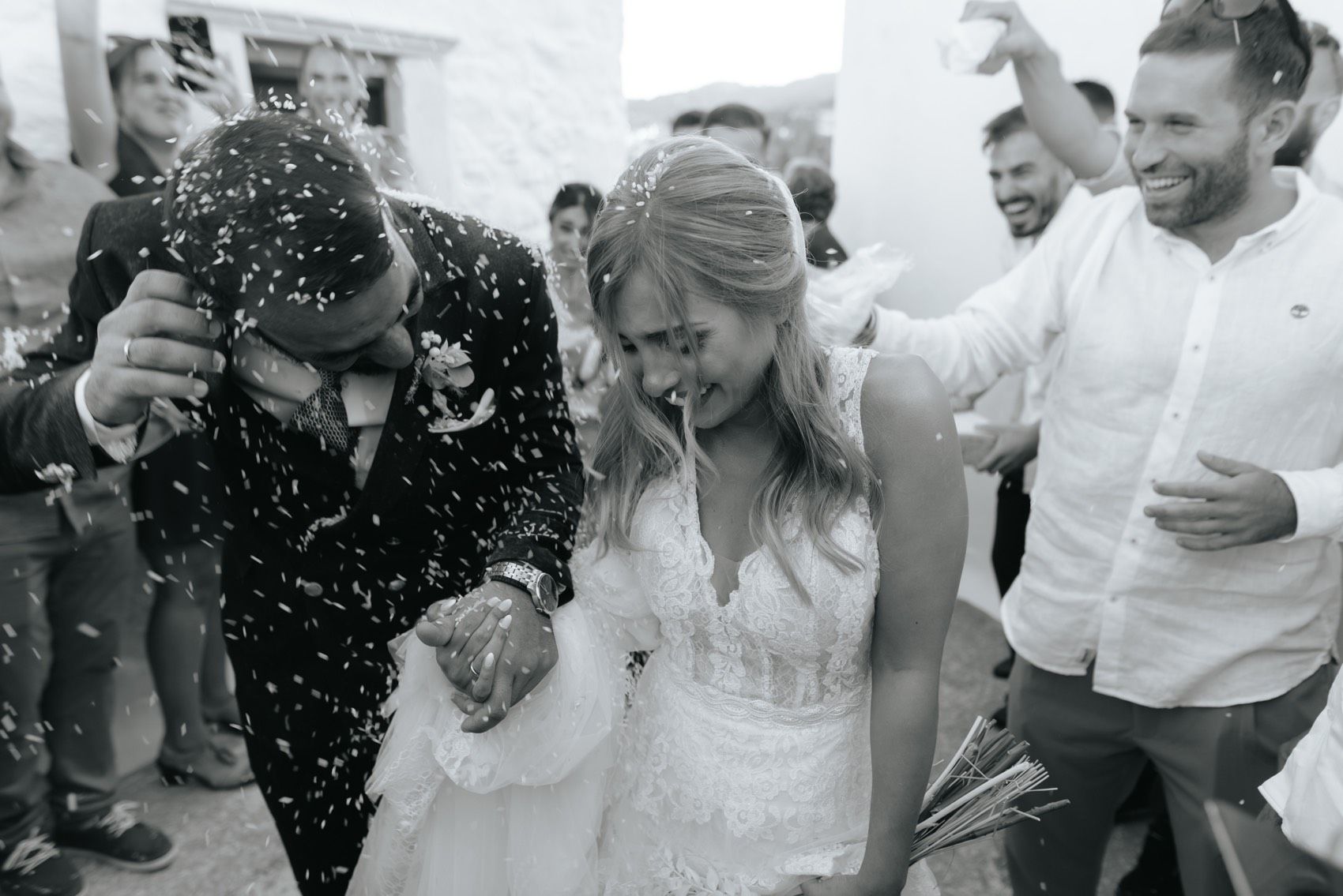Joyful wedding celebration with couple receiving a shower of confetti as guests laugh during outdoor ceremony.