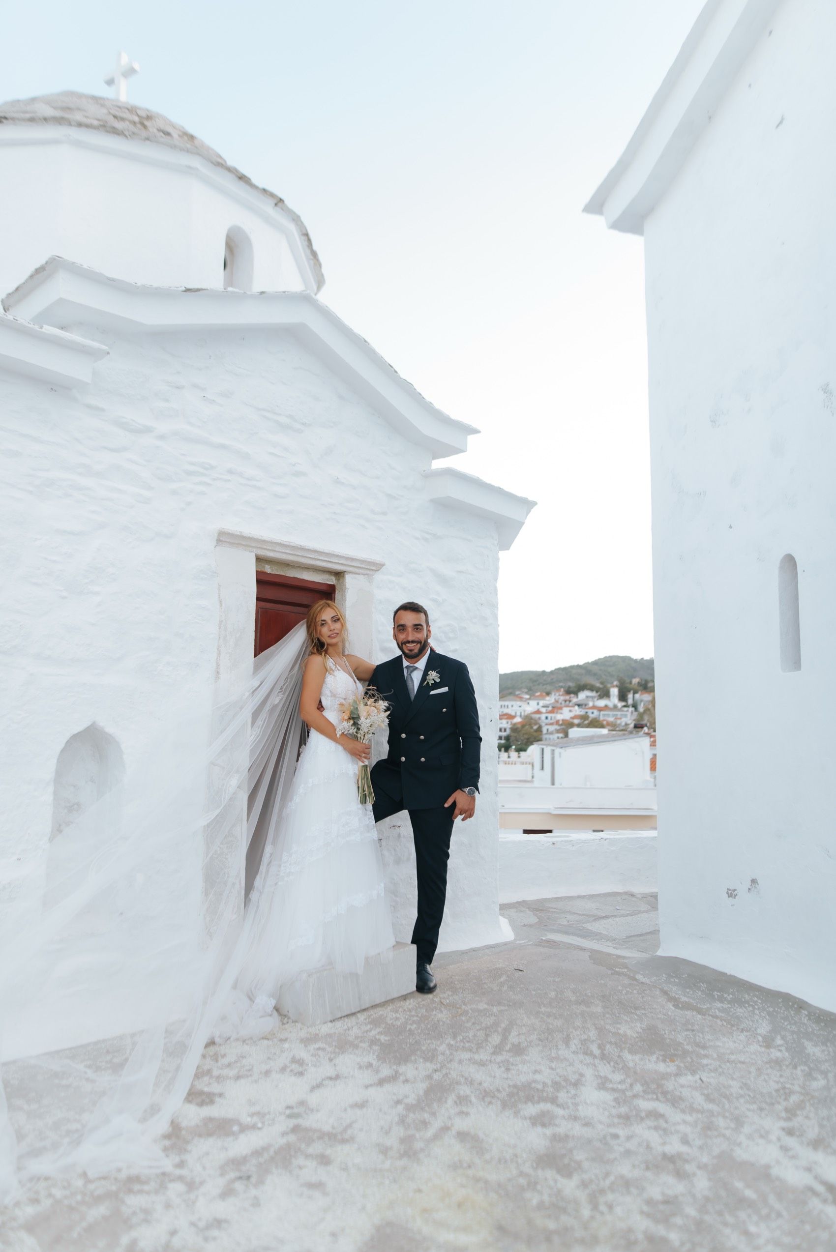 A wedding couple poses outside a traditional whitewashed Greek Orthodox church with domed roof in Santorini.