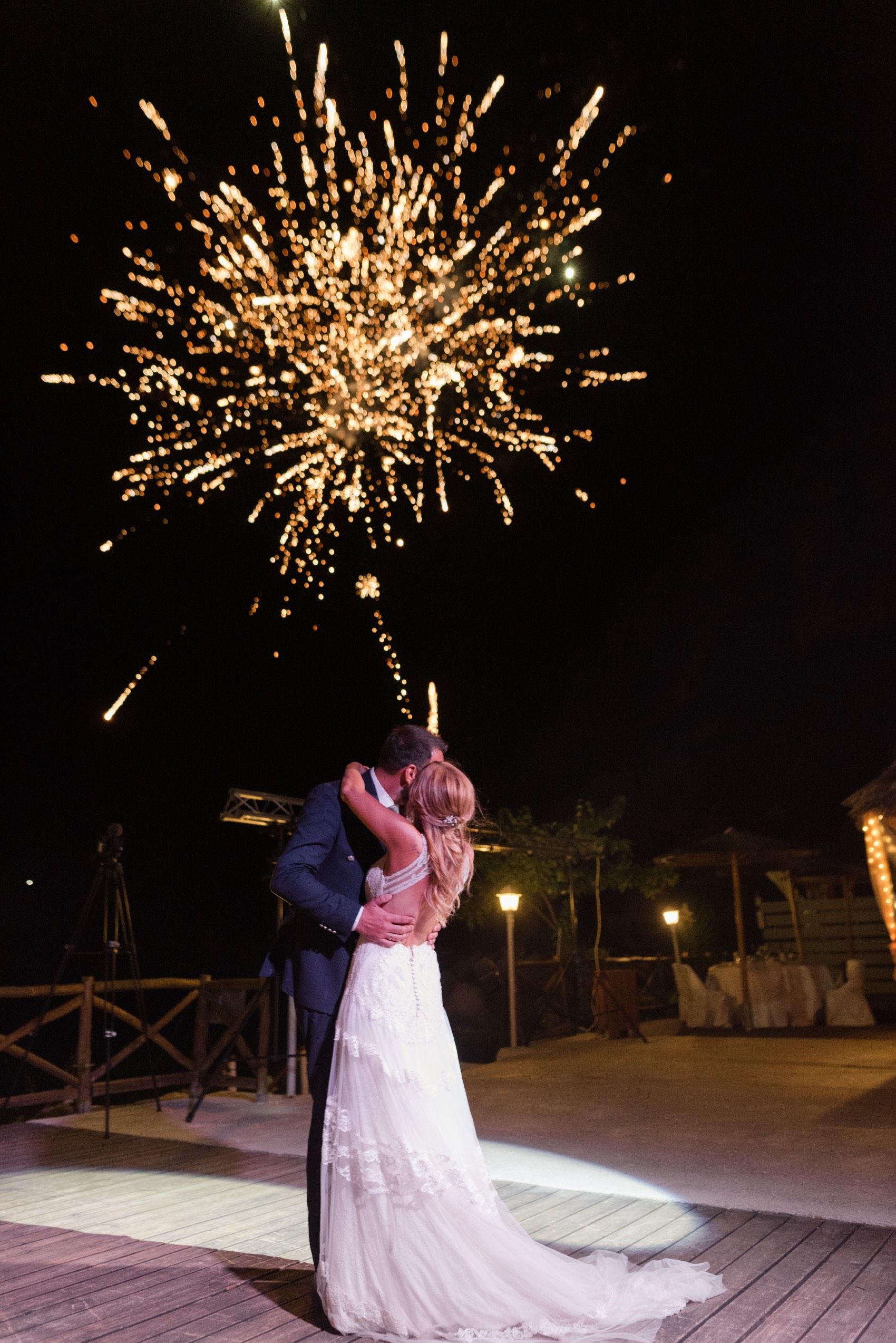 A couple shares a romantic moment under bursting fireworks on a wooden deck during a nighttime celebration.
