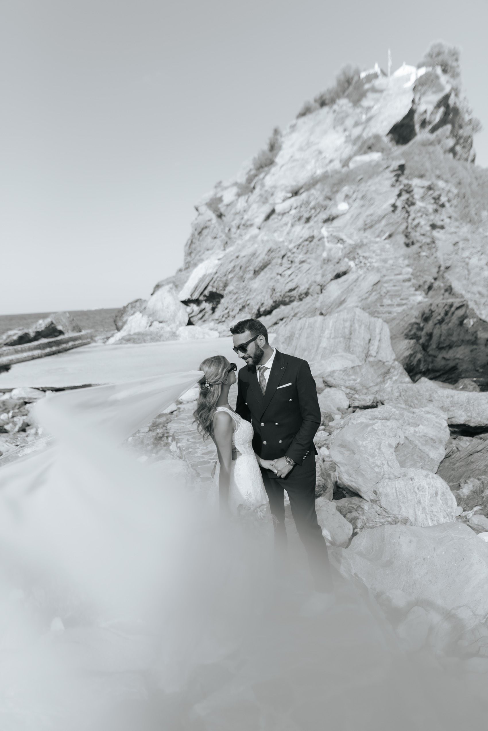 A romantic black and white coastal photograph with a couple embracing near dramatic rocky cliffs by the ocean.