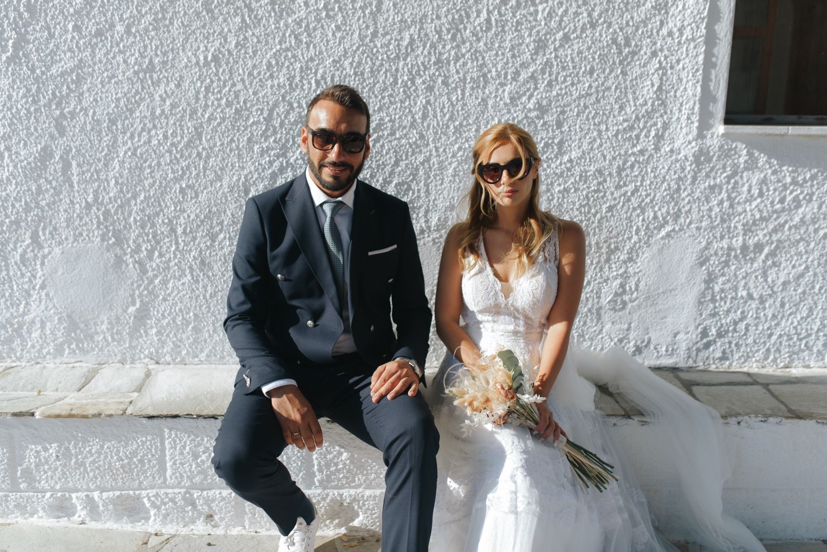 A couple in formal wedding attire sits together on white steps against a white wall on a sunny day.