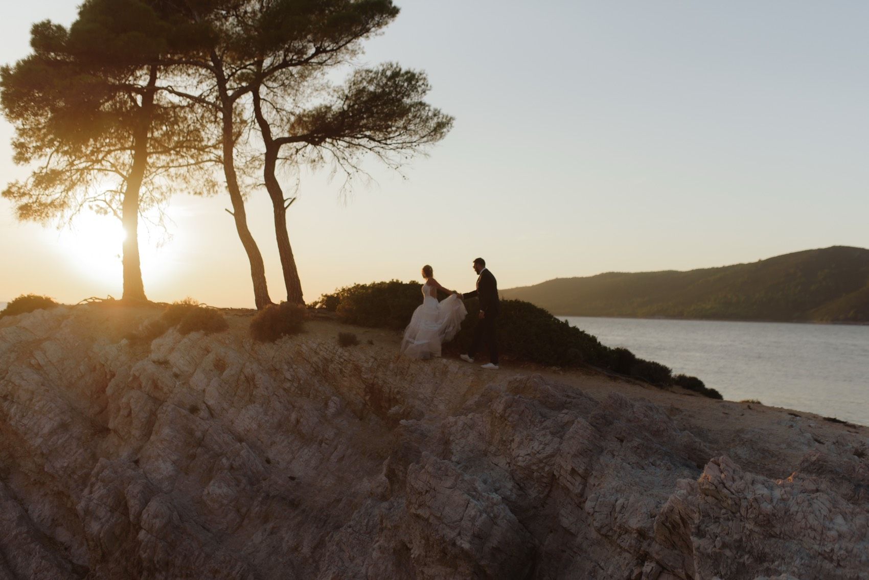 A romantic silhouette of figures beneath pine trees on a rocky cliff overlooking a bay at golden sunset.
