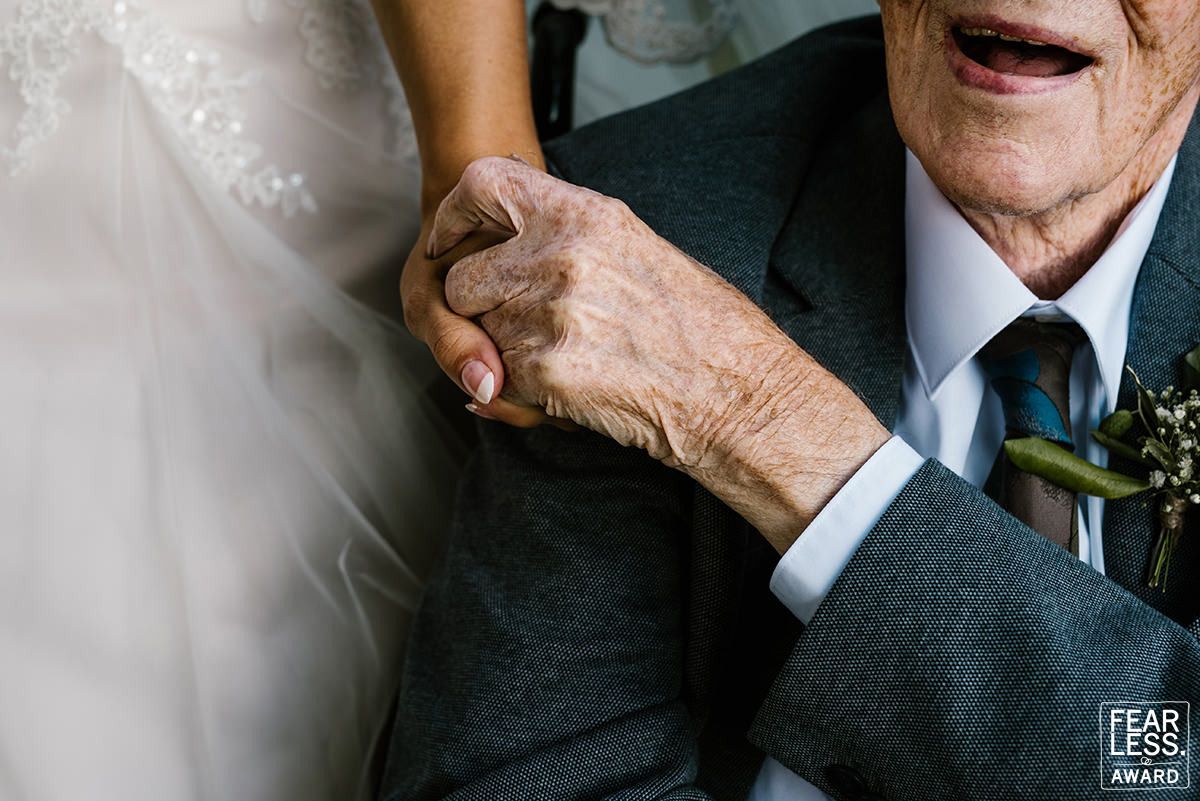 Close-up photo showing hands clasped together, with one wearing a dark suit jacket and boutonniere at a wedding celebration.