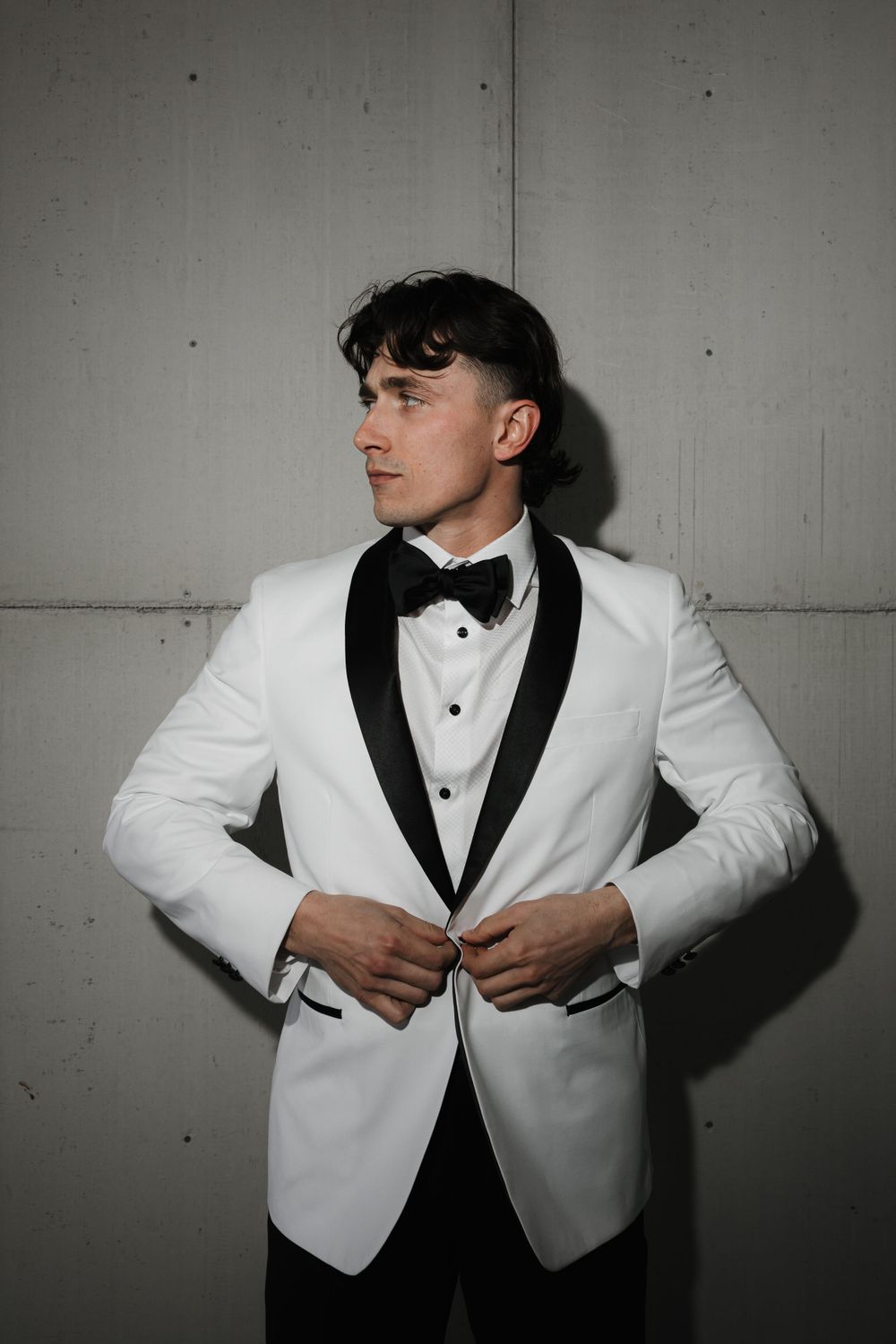 Groom in white tuxedo jacket with black bow tie poses against neutral wall.