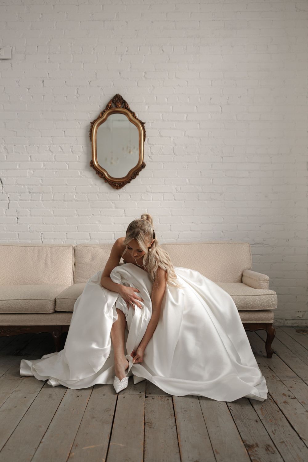 Bride in flowing white gown sitting elegantly on beige couch beneath vintage mirror.