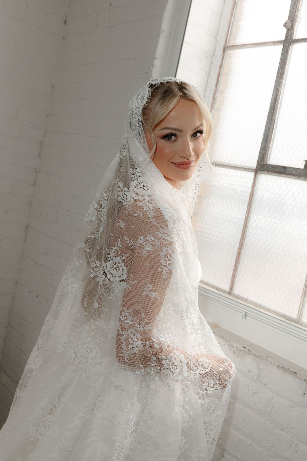 Bride wearing intricate lace veil poses by window in classic wedding portrait.