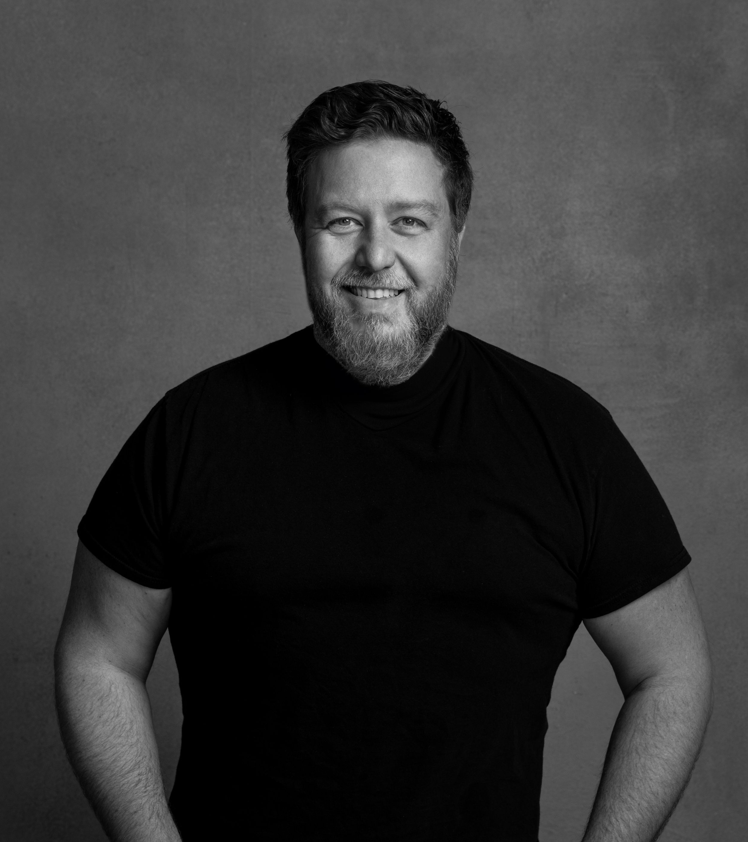 Black and white portrait of a smiling man in a black t-shirt against a gray background.