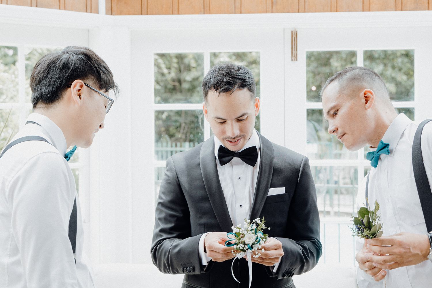Three people in formal attire stand together looking at small floral boutonnieres captured by Apertured Media Auckland Wedding Photographer.