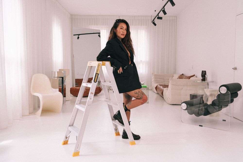 A person poses leaning against a white ladder in a clean, minimalist studio space wearing a black dress and heels.