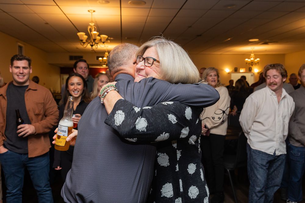 Guests sharing warm embraces at an indoor celebration event with others looking on.