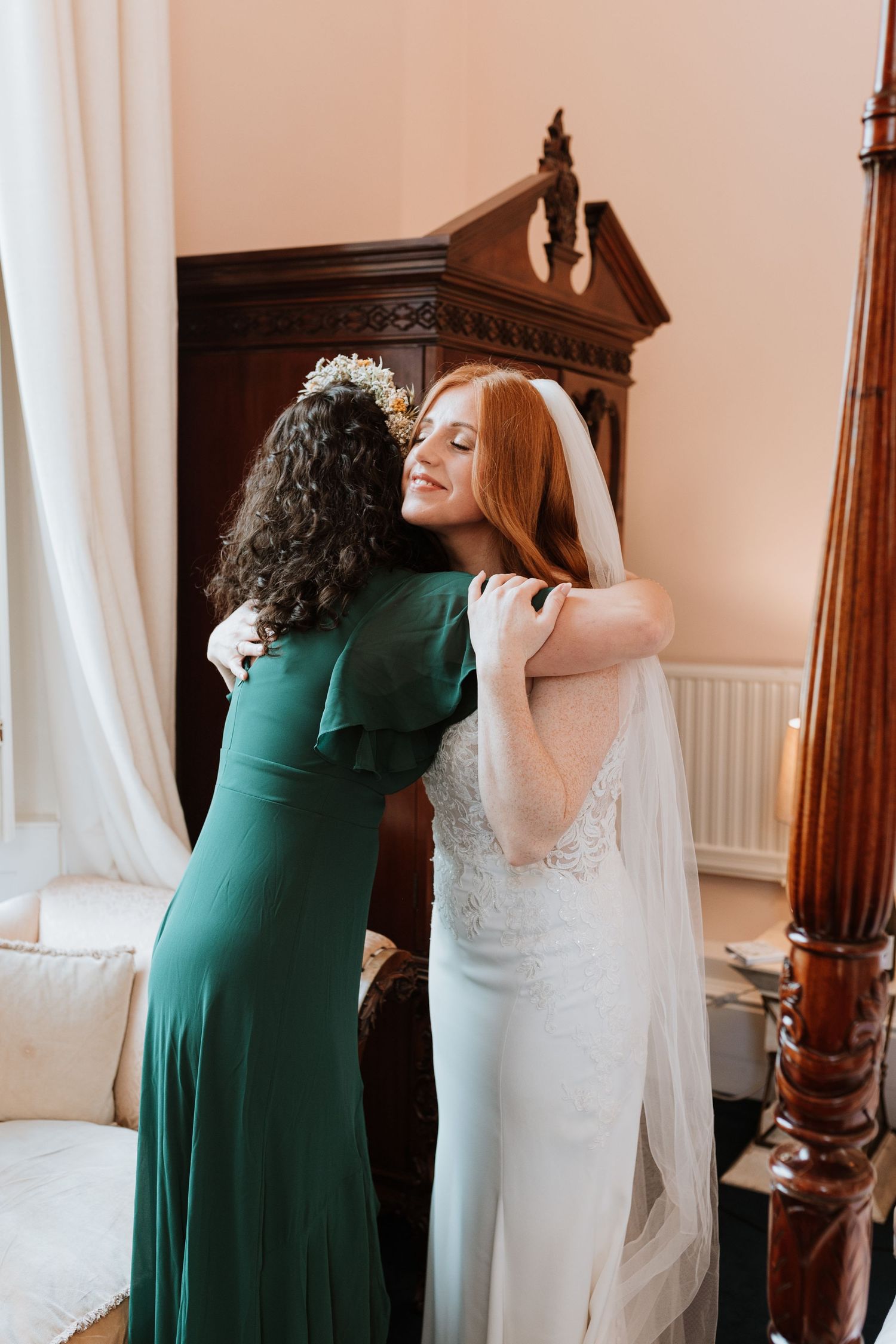 Two women embrace in a tender moment, one wearing an emerald green dress and one in a white wedding gown with veil in Shrewsbury