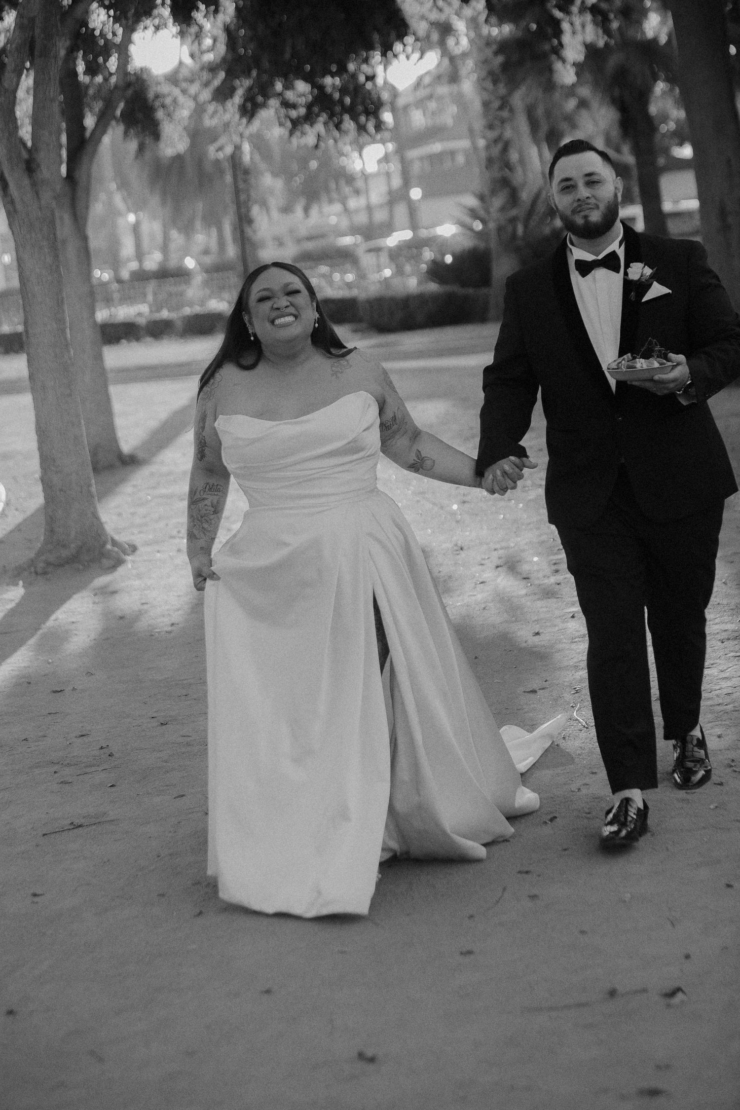 A couple walks together outdoors on their wedding day, with the bride in a white gown and groom in a tuxedo.