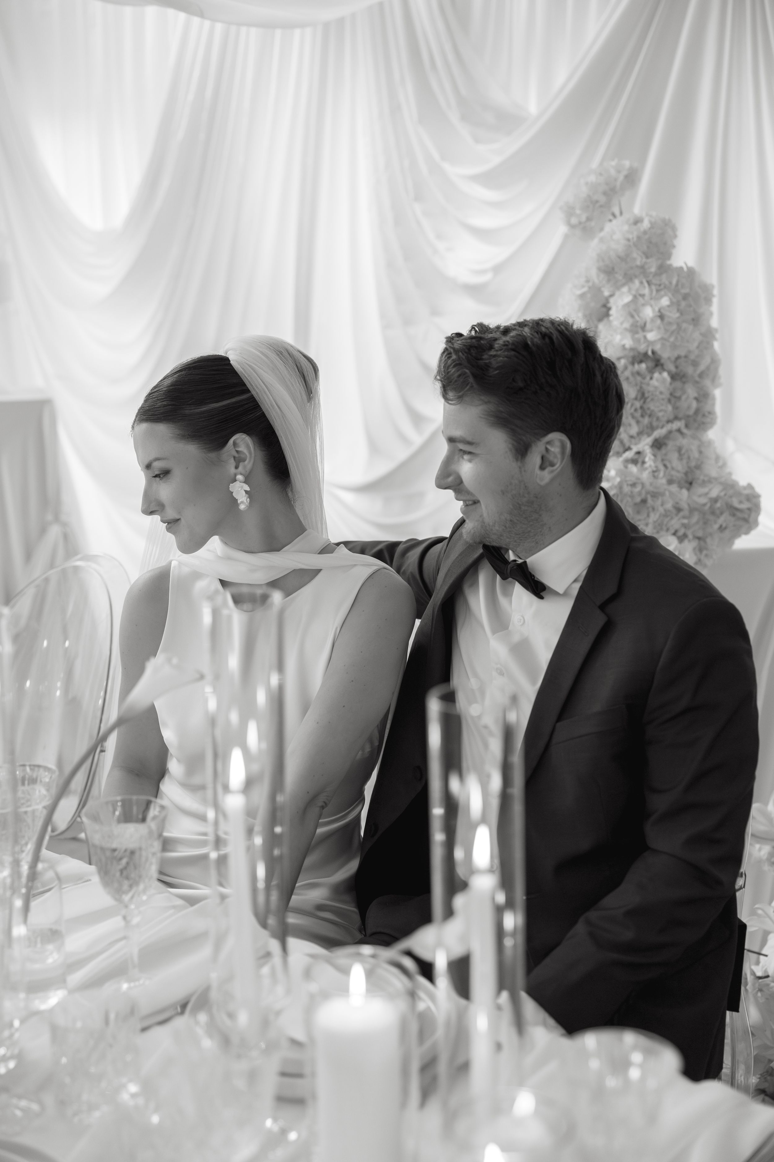 A black and white wedding reception photo showing a candlelit table with a couple seated at their elegant celebration.
