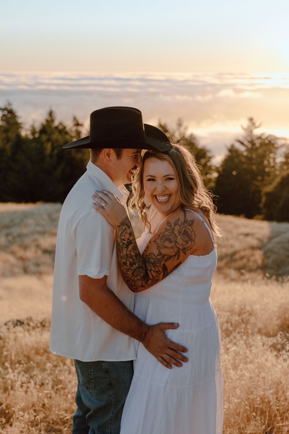 A couple embraces in a golden sunset with evergreen trees in the background, wearing white clothing in a grassy field.