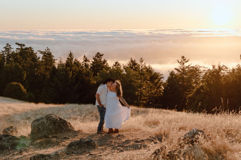 Silhouetted couple stands on golden hillside overlooking fog-filled valley at sunset with evergreen forest backdrop.