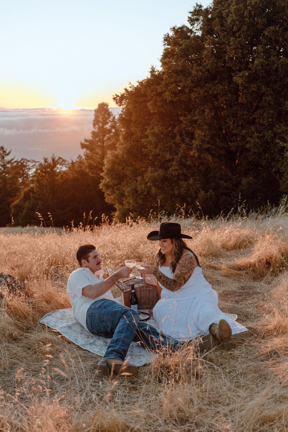 A couple enjoys a romantic sunset picnic in a golden field with wine glasses and mountain views.